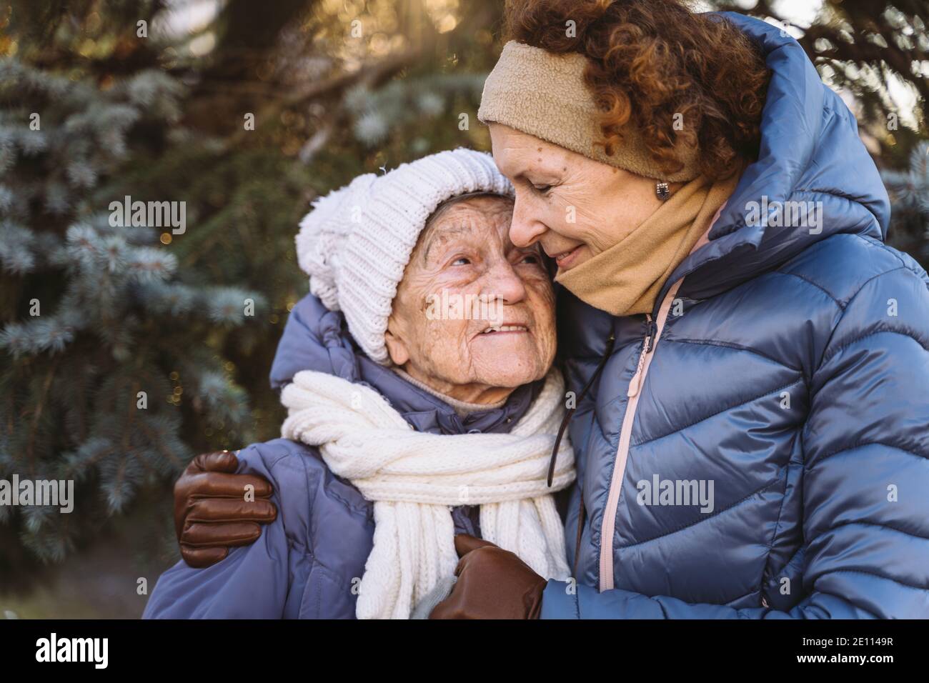Caucasian women senior mother and mature daughter in winter garden on background fir tree hug each other face to face, smile, spend time with old pare Stock Photo
