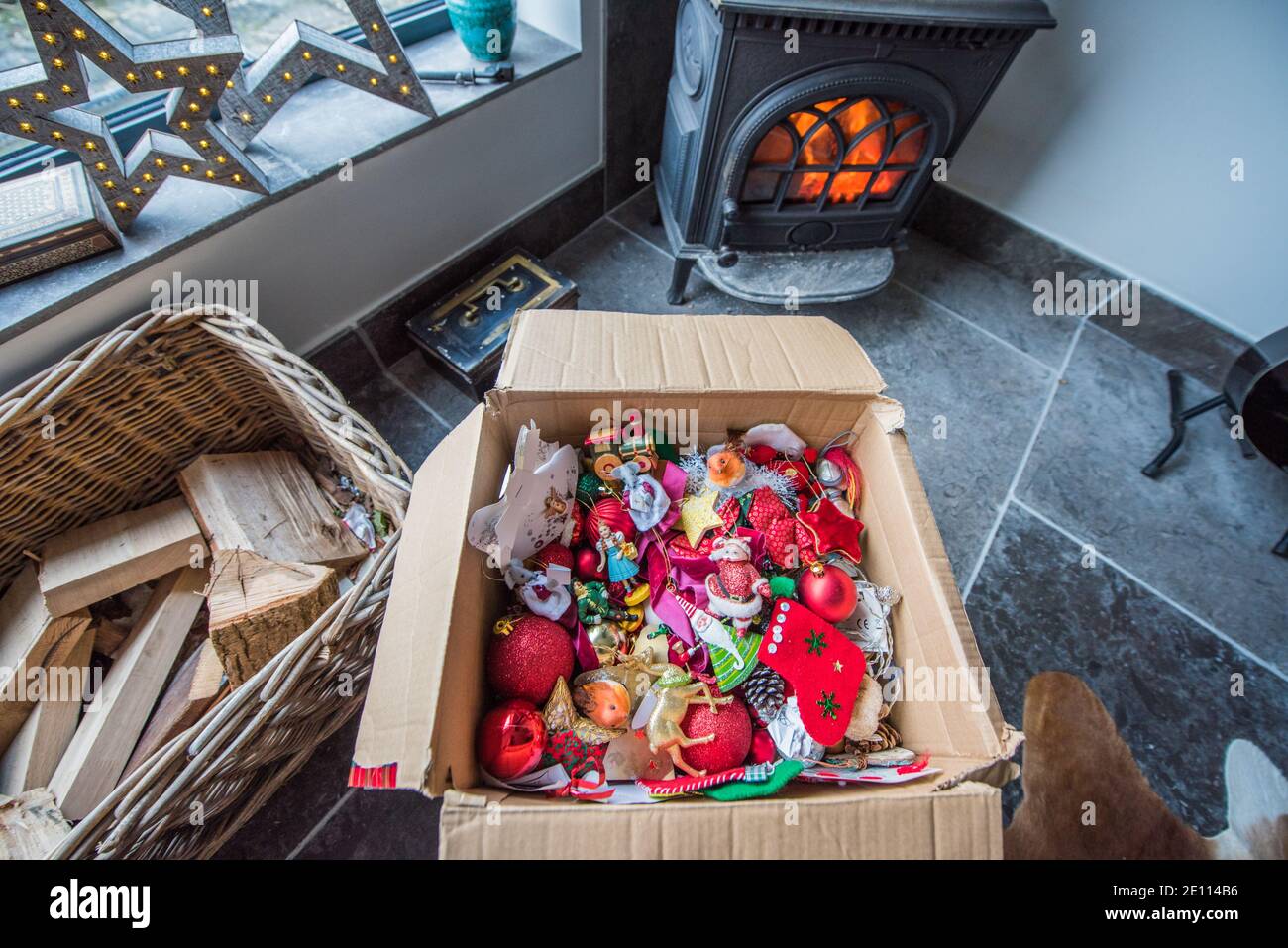 Brown cardboard box with a jumble of family colourful Christmas tree decorations infront of a log burner on a stone floor being packed away Stock Photo