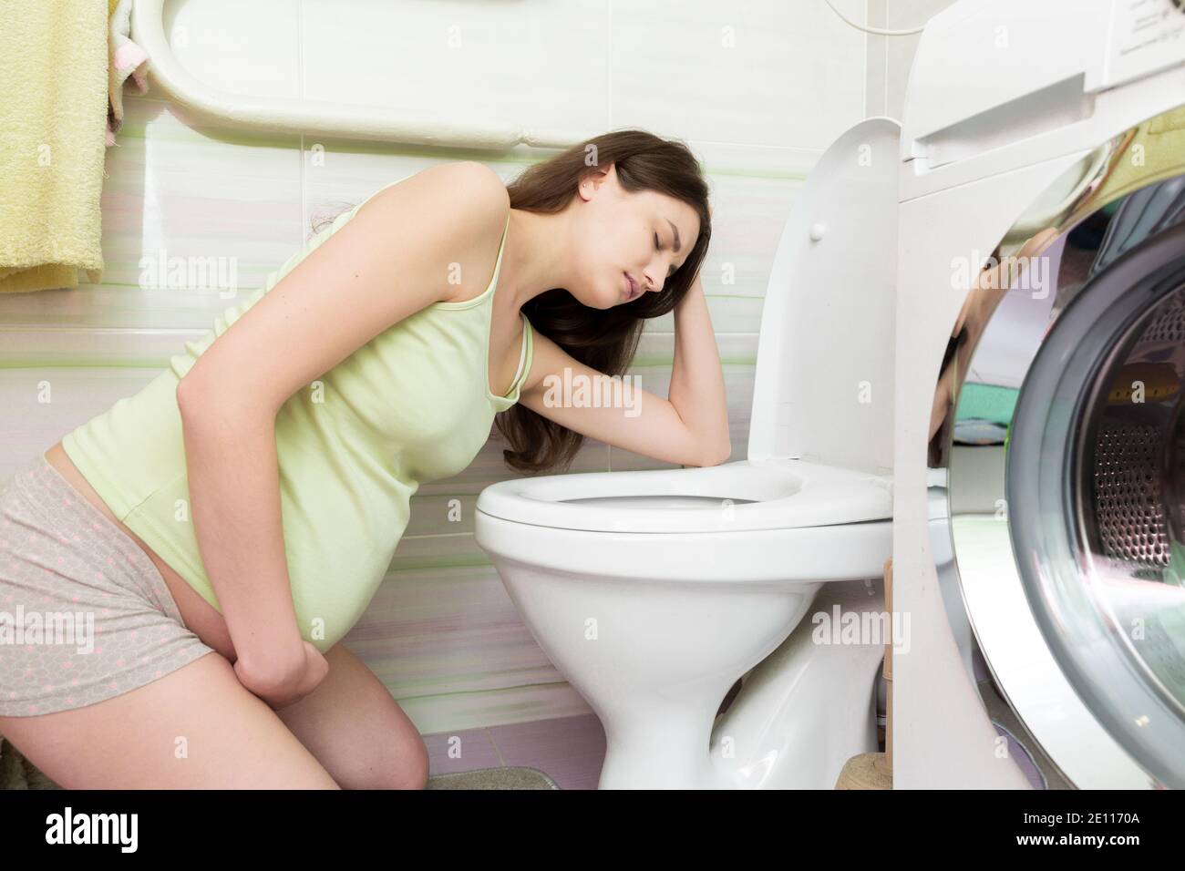 Sick young woman with pain in stomach is vomiting in toilet sitting on the floor at home. Young woman vomiting into the toilet bowl in bathroom. Stock Photo