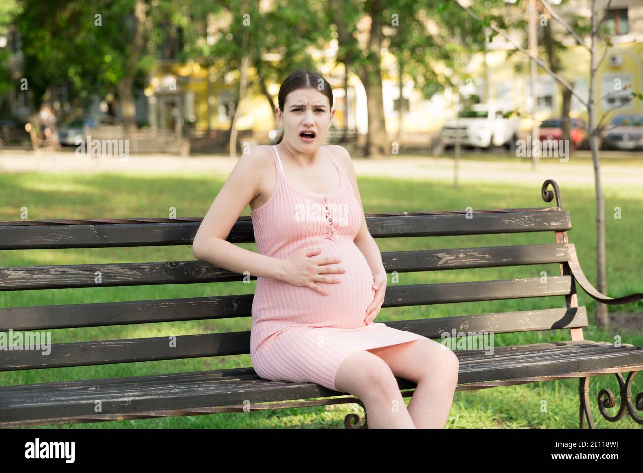 A pregnant young woman on a bench sitting in the park and holds on to the stomach which hurts. Pregnancy and motherhood. Healthy lifestyle. Stock Photo