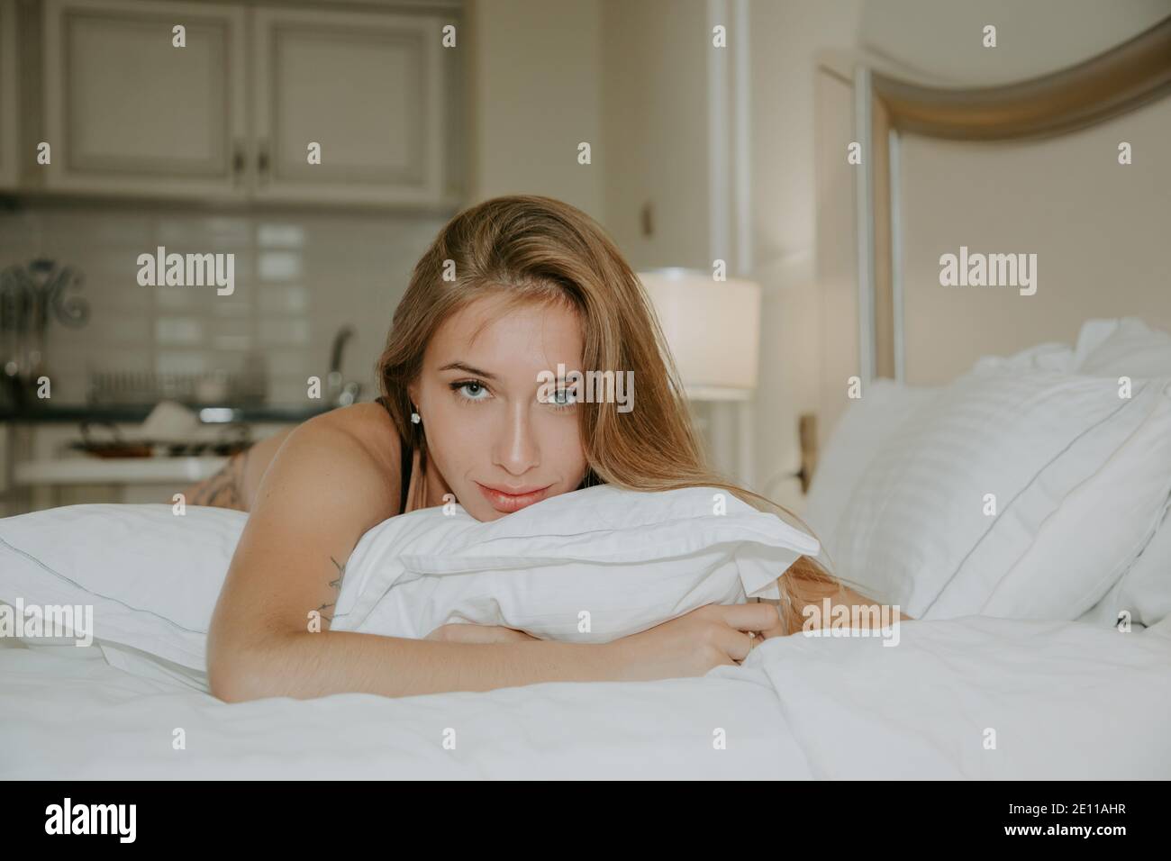Portrait of a pretty girl with long hair lying on a white pillow on her bed; she looks natural, She is looking intensely at us. Kitchen in background Stock Photo