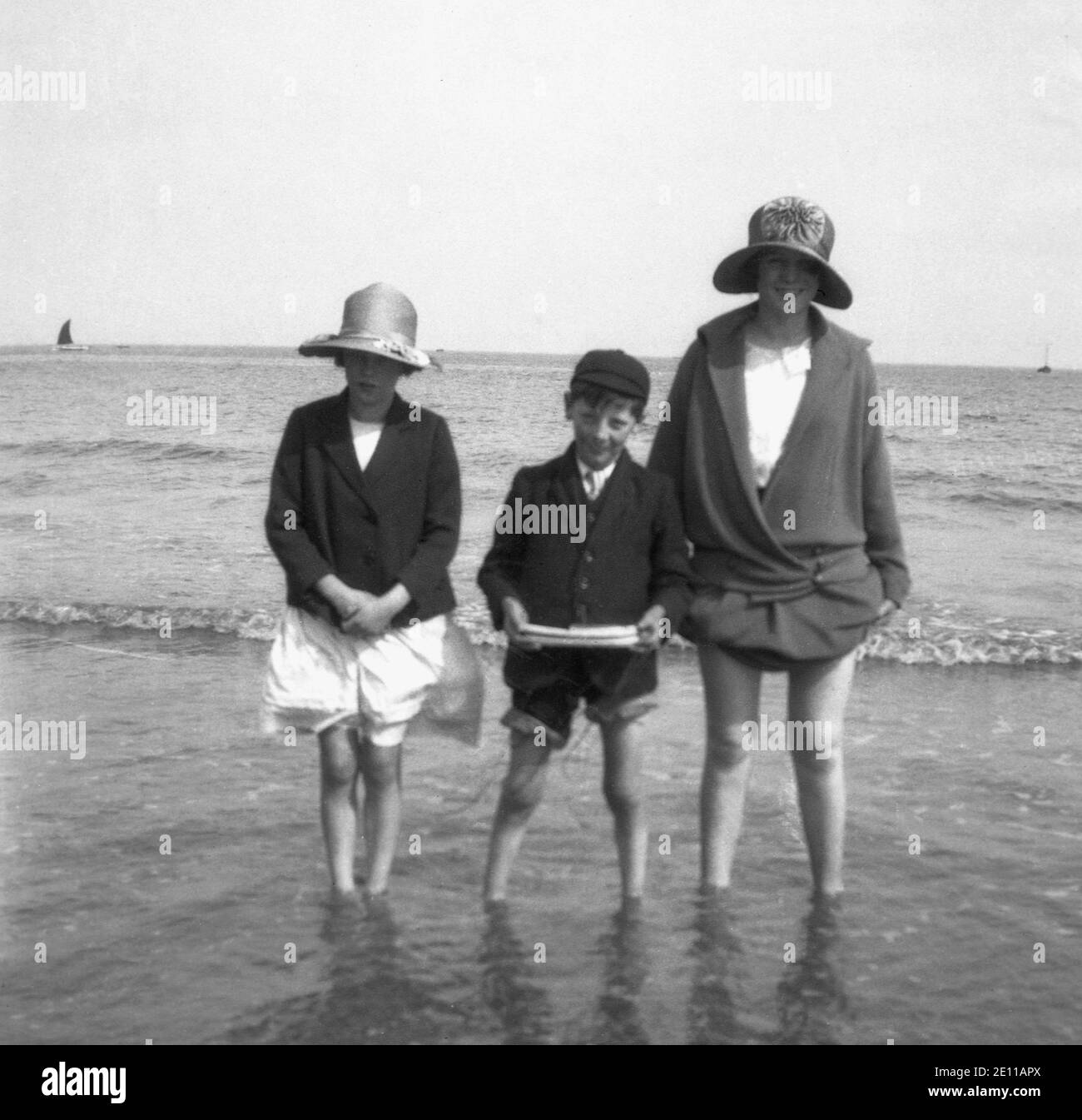 1930s, ...oh its nice to be beside the sea... a mother with her two children in their school blazers doing what the British do at the seaside, have a paddle with the trousers and skirts rolled up and showing off their 'knobbly' knees! Stock Photo
