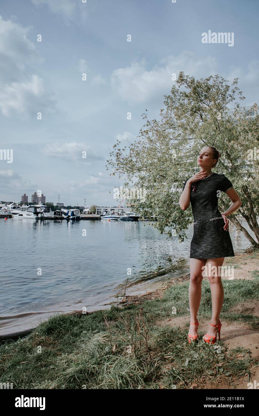 Pretty blonde girl stands in park near the water with boats in the background; she is wearing a stylish short dark dress and high heels Stock Photo