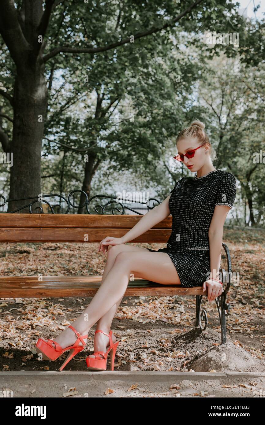 Pretty blonde girl sits on a bench in a park; she is wearing a stylish short dark dress, red sunglasses and high heels Stock Photo
