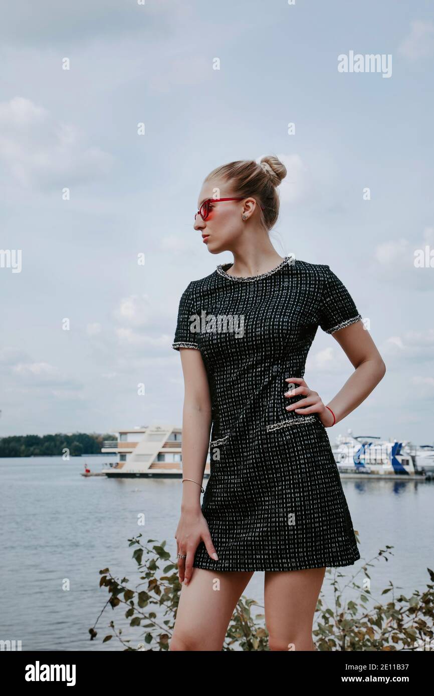 Pretty blonde girl stands near the water with boats in the background; she is wearing a stylish short dark dress and red sunglasses. Stock Photo