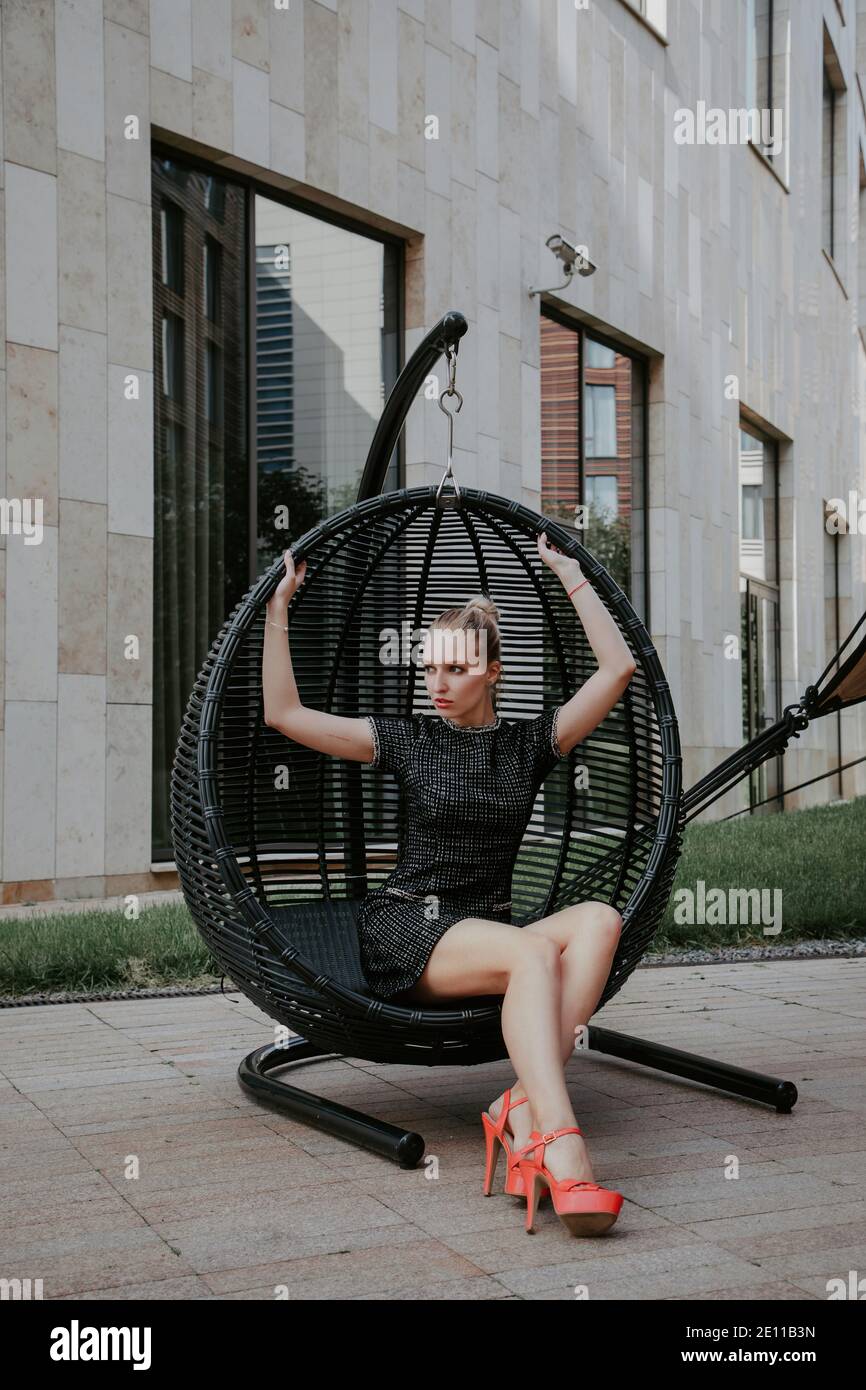 Pretty blonde girl sits in a hanging chair in an modern urban environment; she is wearing a  stylish short dark dress and high heels Stock Photo