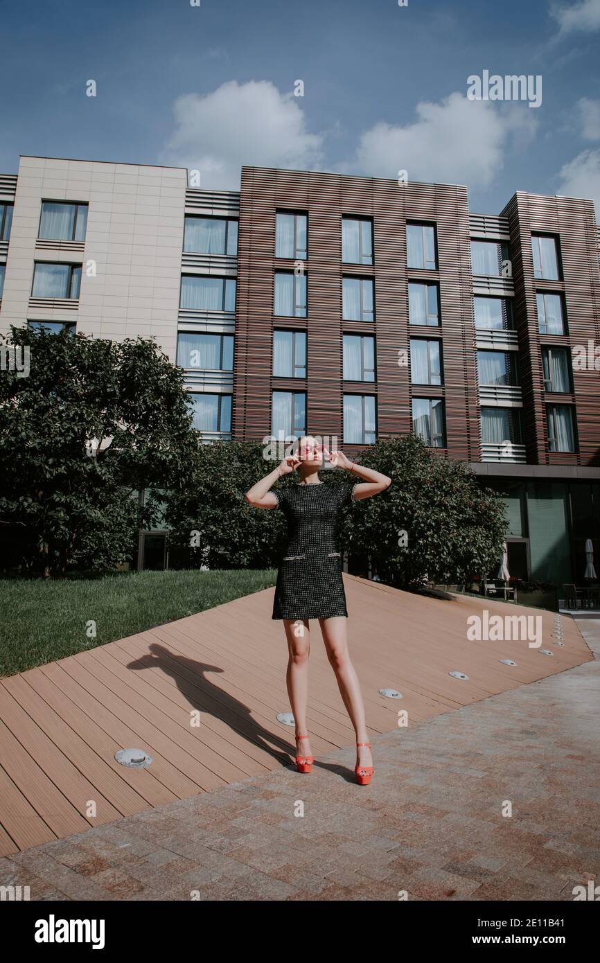 Pretty blonde girl stands in modern urban environment; she is wearing a stylish short dark dress & high heels; she looks up holding her red sunglasses Stock Photo