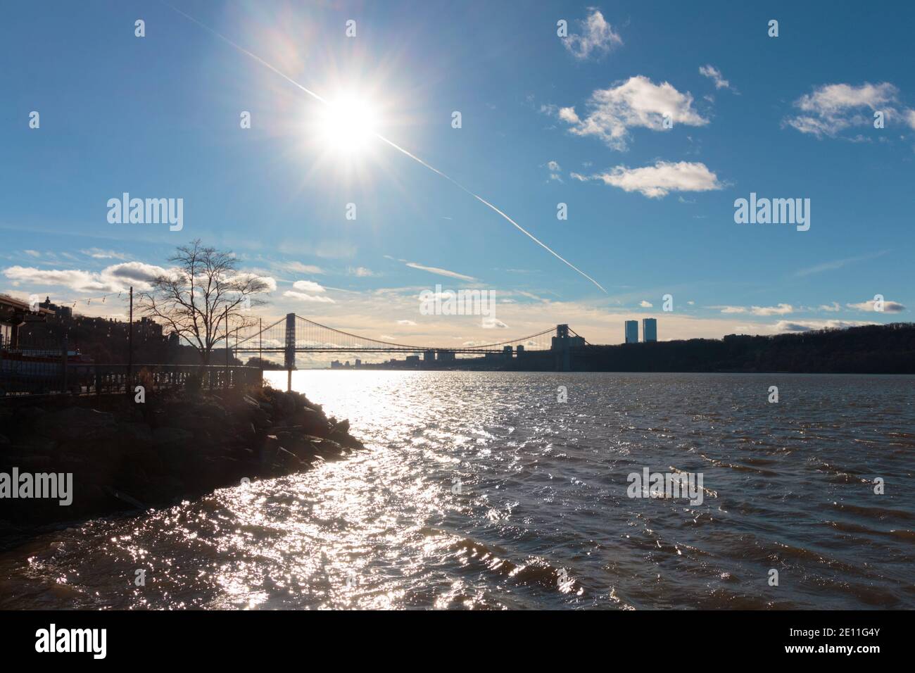 a bright white winter sun shines over the Hudson River with an airplane contrail passing through it, the George Washington Bridge  in the far distance Stock Photo