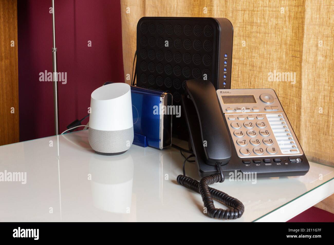 home office set up with a voice activated, virtual smart assistant, a landline telephone, and two routers on a white desk with  a burgundy and yellow Stock Photo