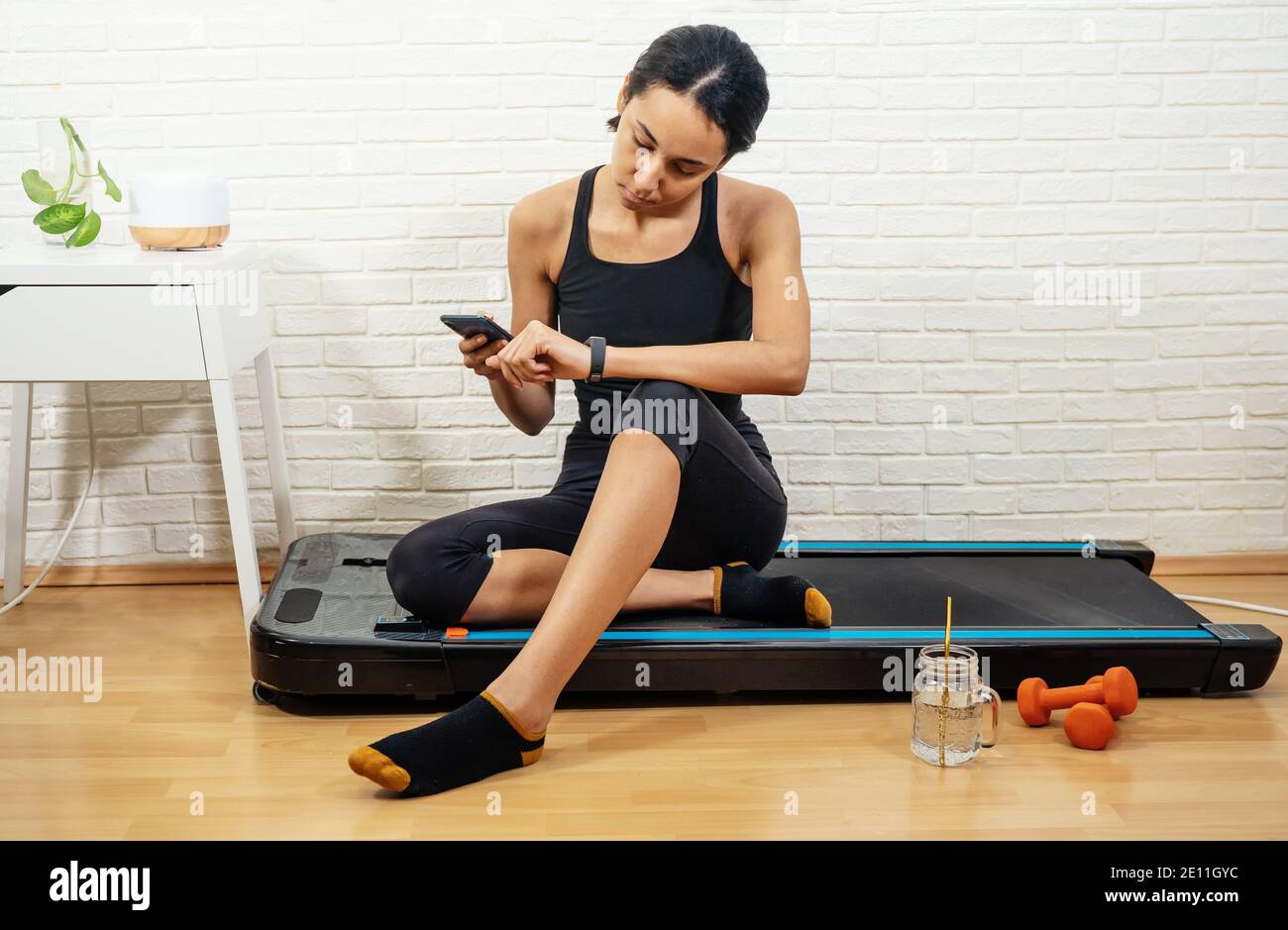 Young mixed-raced woman measures activity control with smartphone after workout at home Stock Photo