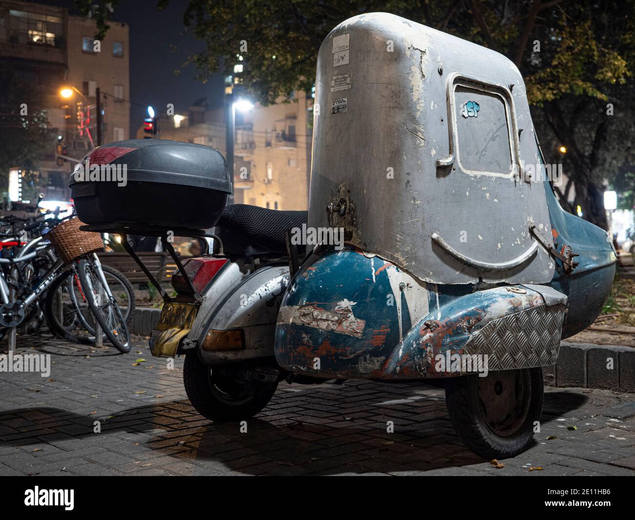 OLD CLASSIC VESPA SCOOTER WITH COVERED SIDECAE SEAT PARKING IN URBAN PARKING LOCATION AT NIGHT. THE SIDE CAR HAS WINDOW AND IT LOOKS LIKE SMILING FACE. Stock Photo