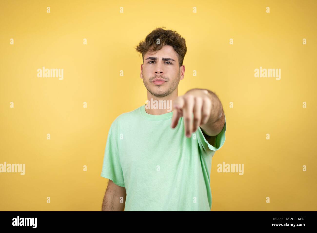 Handsome man wearing a green casual t-shirt over yellow background serious pointing to the front with finger Stock Photo