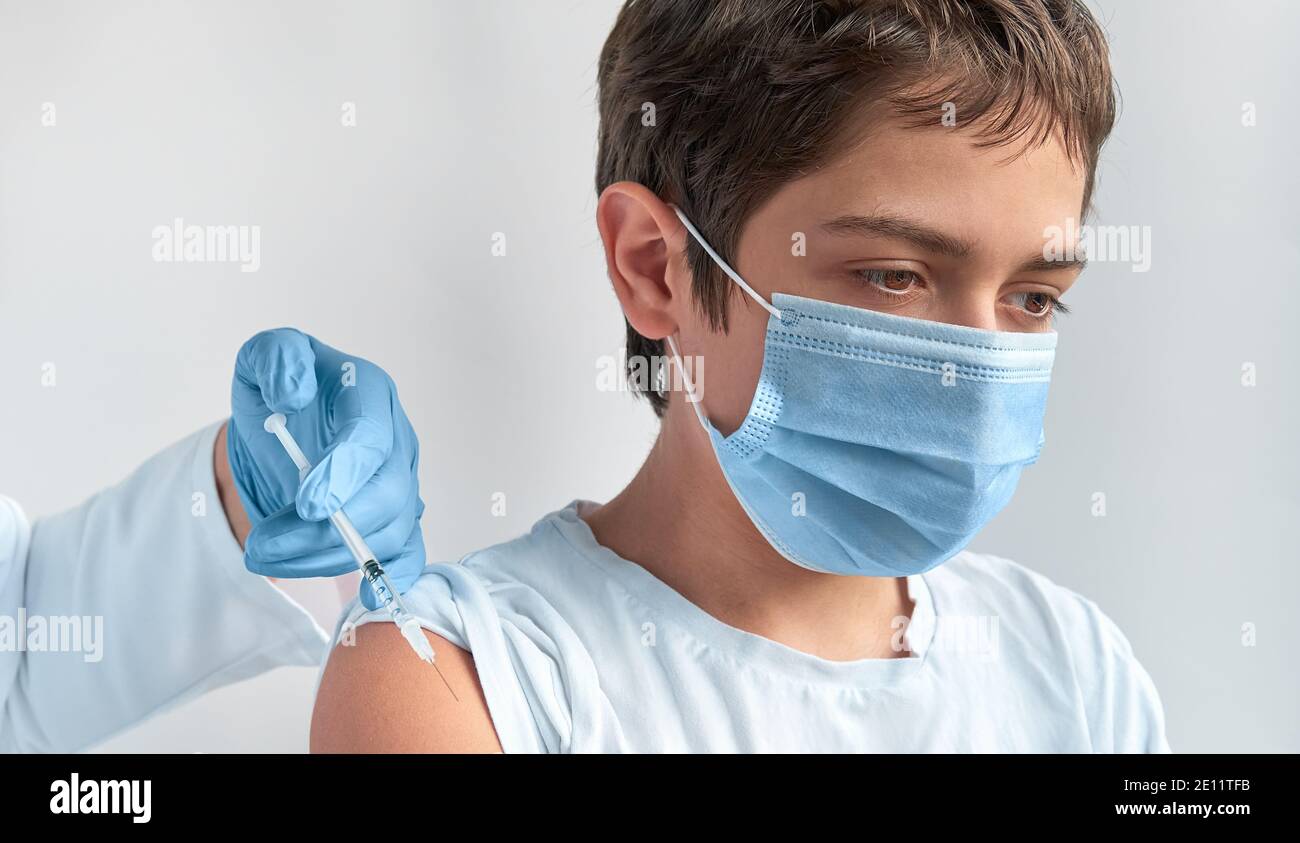 Covid 19, flu or measles vaccination concept. Caucasian teenage boy in face mask, and hand with syringe. Medic, doctor or nurse vaccinates teenage Stock Photo