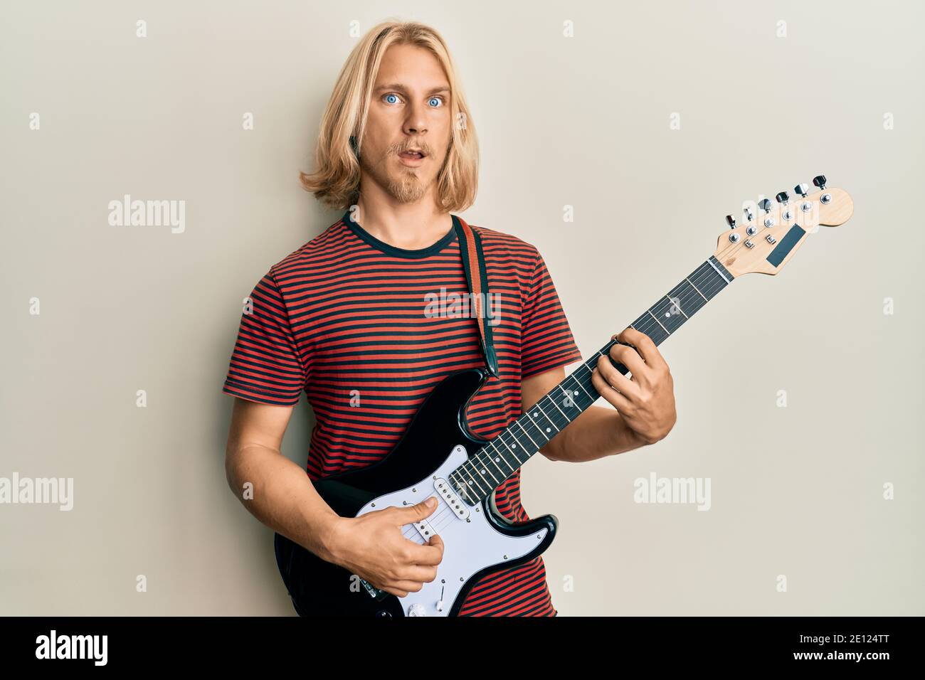 Caucasian young man with long hair playing electric guitar in shock face,  looking skeptical and sarcastic, surprised with open mouth Stock Photo -  Alamy
