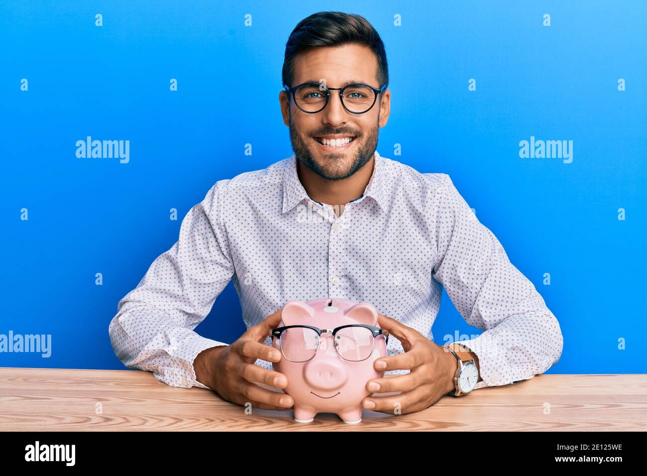 Handsome hispanic man holding piggy bank with glasses smiling with a ...
