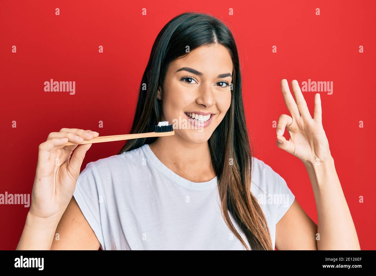 Young brunette woman holding toothbrush with toothpaste doing ok sign ...