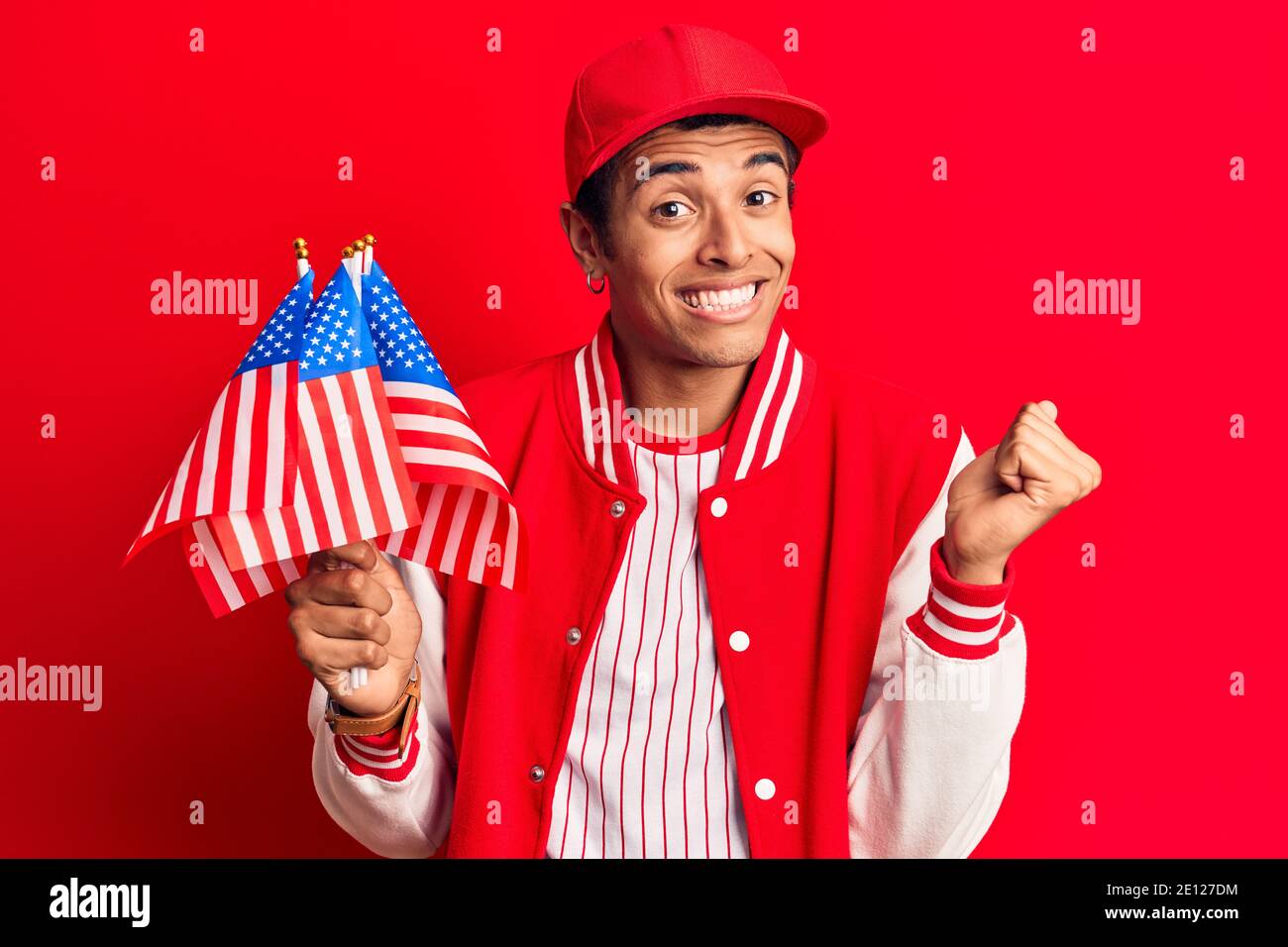 Young african amercian man wearing baseball uniform holding america flags screaming proud, celebrating victory and success very excited with raised ar Stock Photo