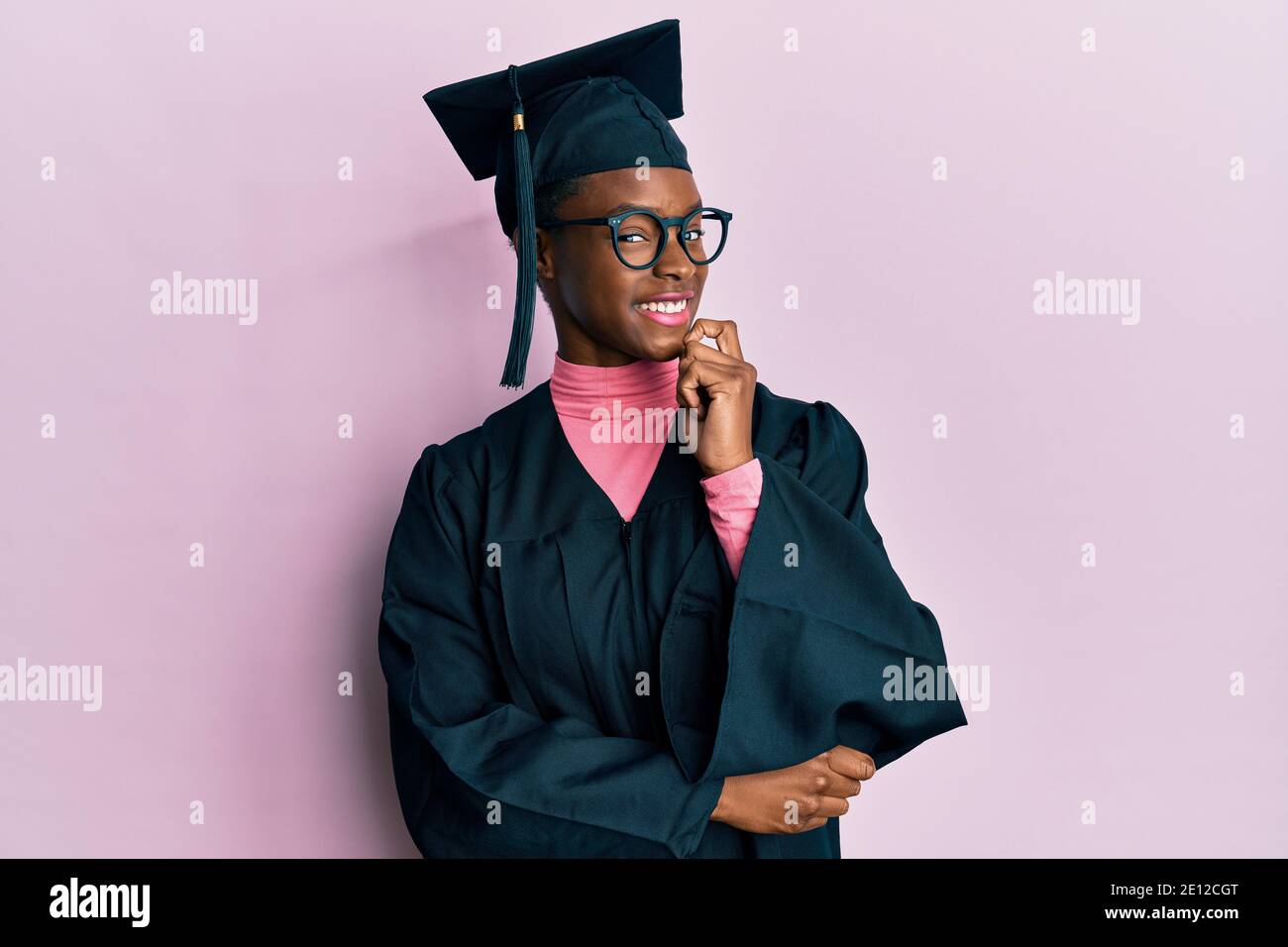 Young african american girl wearing graduation cap and ceremony robe looking confident at the camera smiling with crossed arms and hand raised on chin Stock Photo