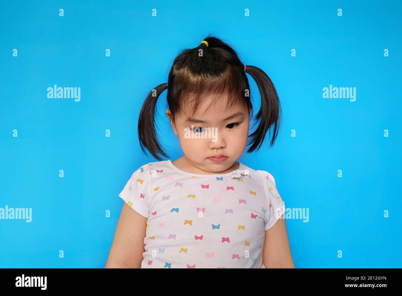 A cute young chubby Asian girl making a sad face, thinking about her punishment. Plain light blue background. Stock Photo
