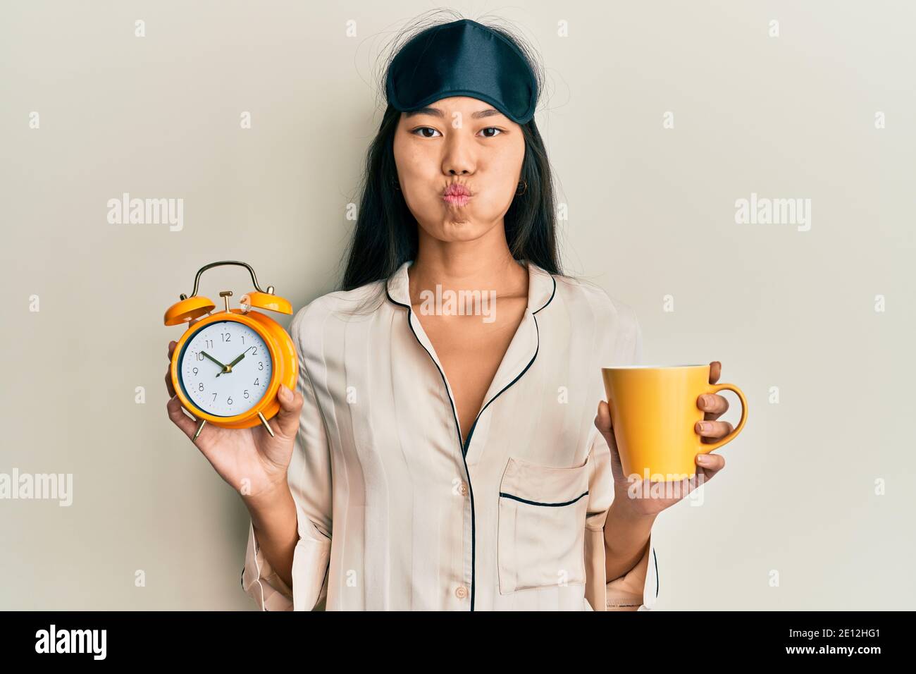 Young chinese woman wearing sleep mask and robe holding alarm clock and cup of coffee puffing cheeks with funny face. mouth inflated with air, catchin Stock Photo