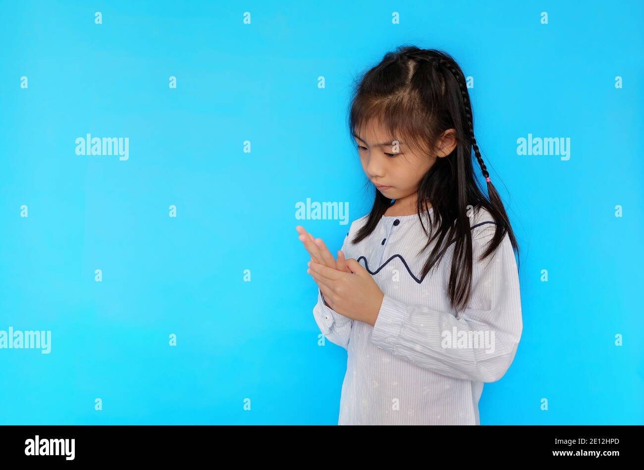 A cute young Asian girl is trying to solve a math problem, using her fingers to count the numbers. Plain light blue background. Stock Photo