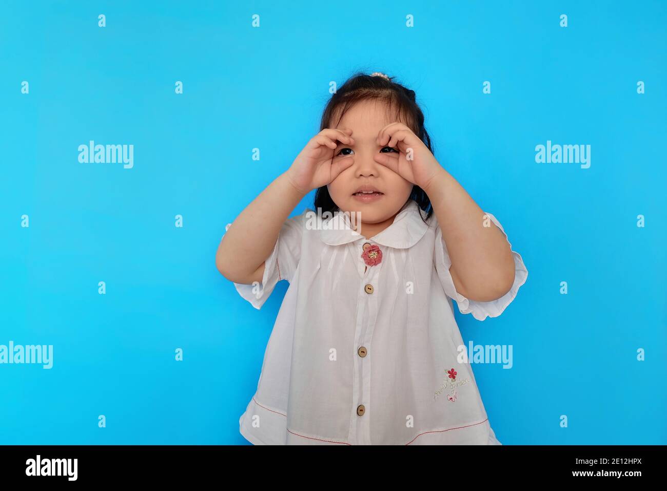 A cute young Asian girl is pretending to be a spy, using her hands as her pretend binoculars, playing and having fun. Plain light blue background. Stock Photo