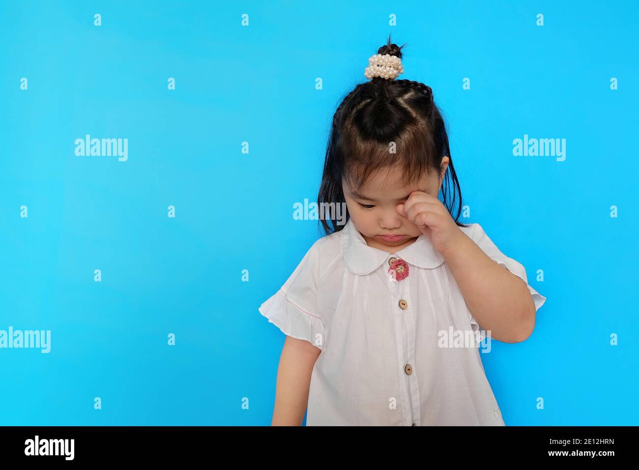 A cute Asian female toddler feeling upset, crying and wiping tears from her eye with her hand. Plain blue background. Stock Photo