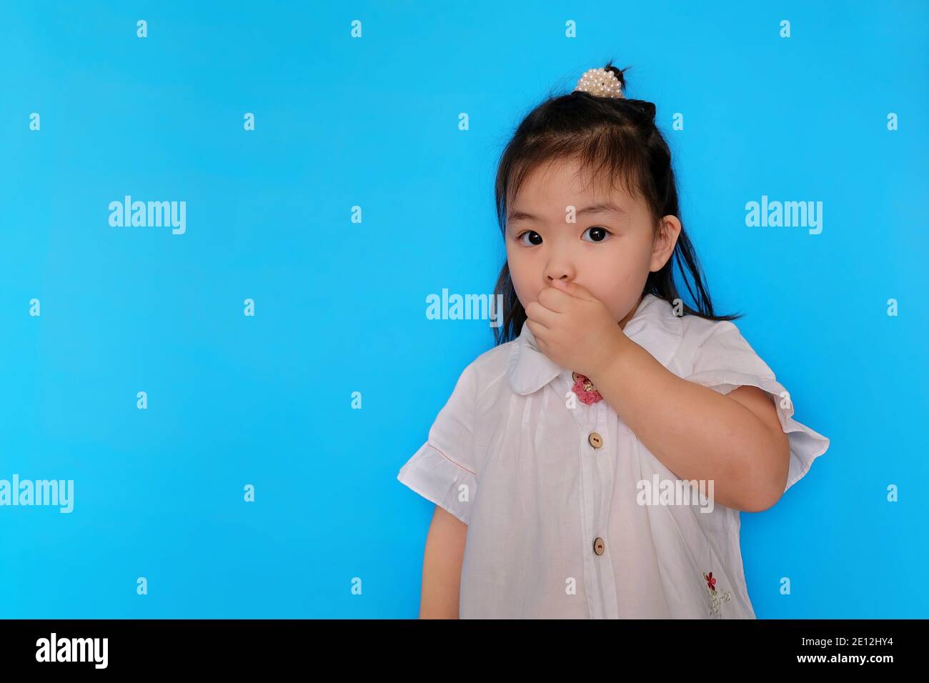A cute young Asian girl is trying to keep a secret, covering her mouth with her hand, acting suspicious. Plain light blue background. Stock Photo