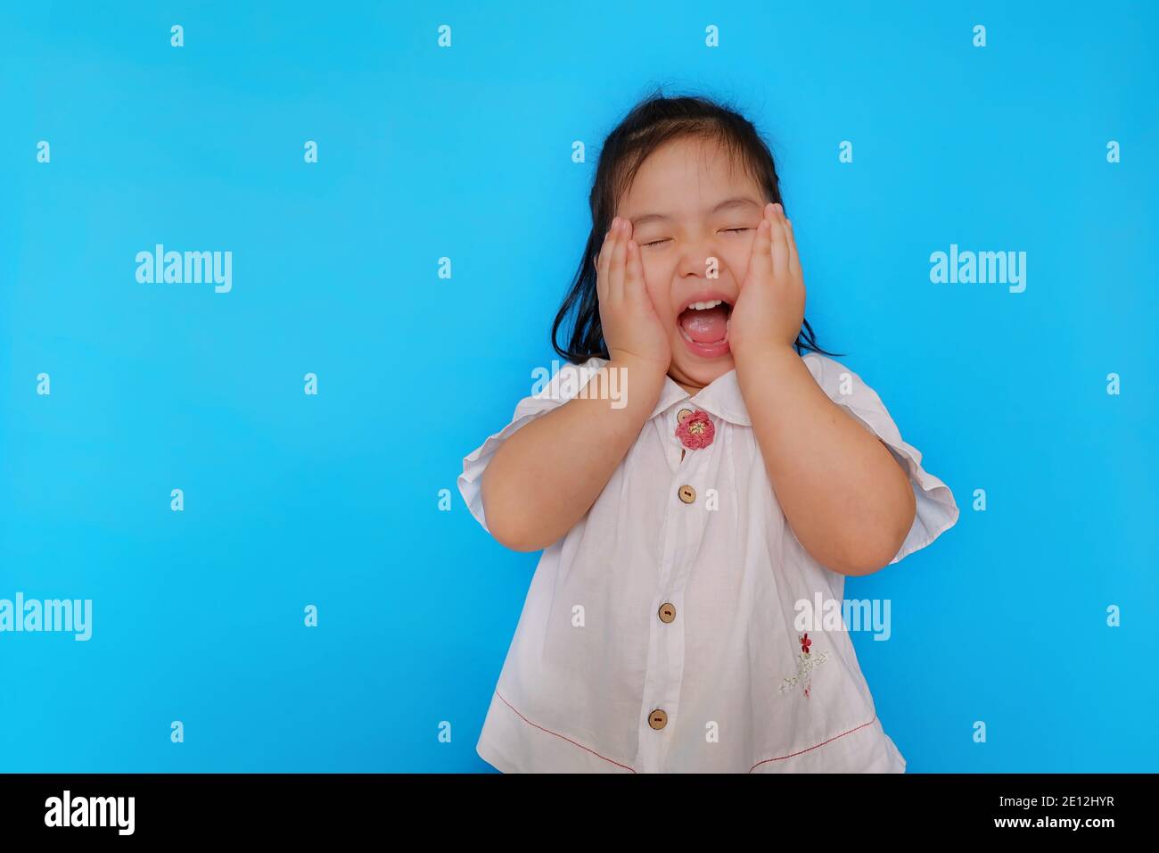 A portrait of a cute Asian girl feeling excited, screaming with her eyes closed and the hands covering her cheek with plain light blue background. Stock Photo
