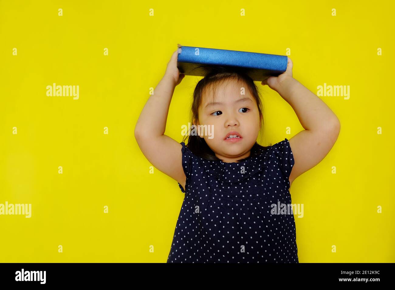 A cute young Asian girl trying to balance a large blue text book on her head as a punishment from her teacher. Bright yellow background. Stock Photo