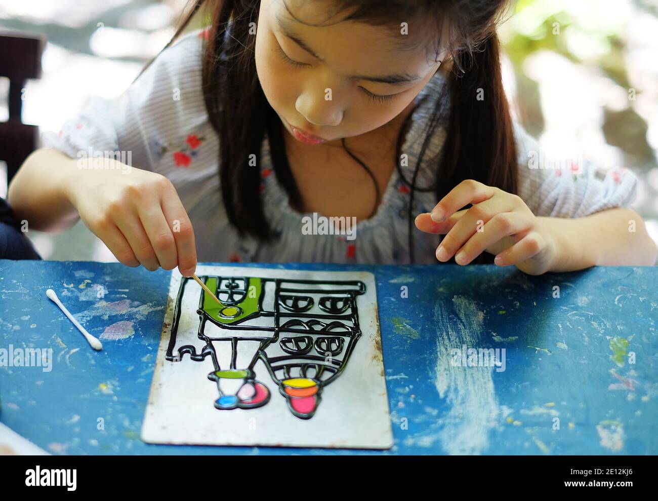A cute Asian girl working on her art project at a daycare center, using colorful acrylic paints to draw a castle. Stock Photo