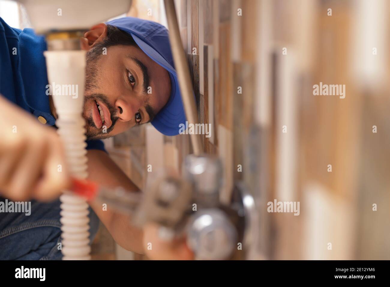 A YOUNG PLUMBER LEANING DOWN AND FIXING LEAKAGE OF A PIPE Stock Photo