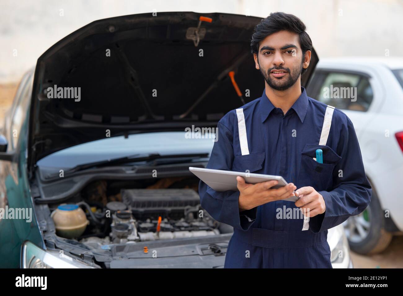 A YOUNG MECHANIC HOLDING DIGITAL TABLET STANDING IN FRONT OF A CAR TO REPAIR Stock Photo