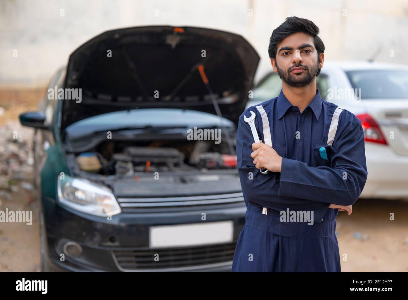 A YOUNG MECHANIC HOLDING TOOLS STANDING IN FRONT OF AN OPEN CAR Stock Photo