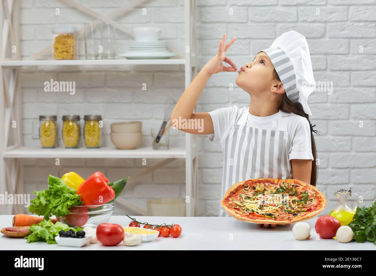 child making tasty delicious gesture by kissing fingers. little girl in chef hat and an apron cooking pizza in the kitchen. Stock Photo