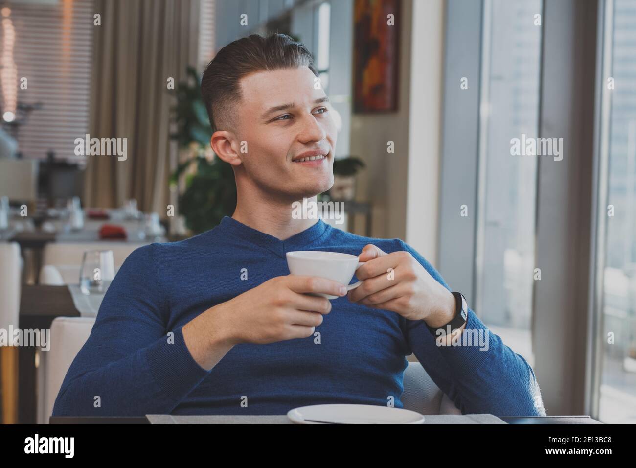 Attractive man smiling, looking in the window, relaxing at the cafe holding cup of delicious coffee. Happy man having breakfast at the restaurant look Stock Photo