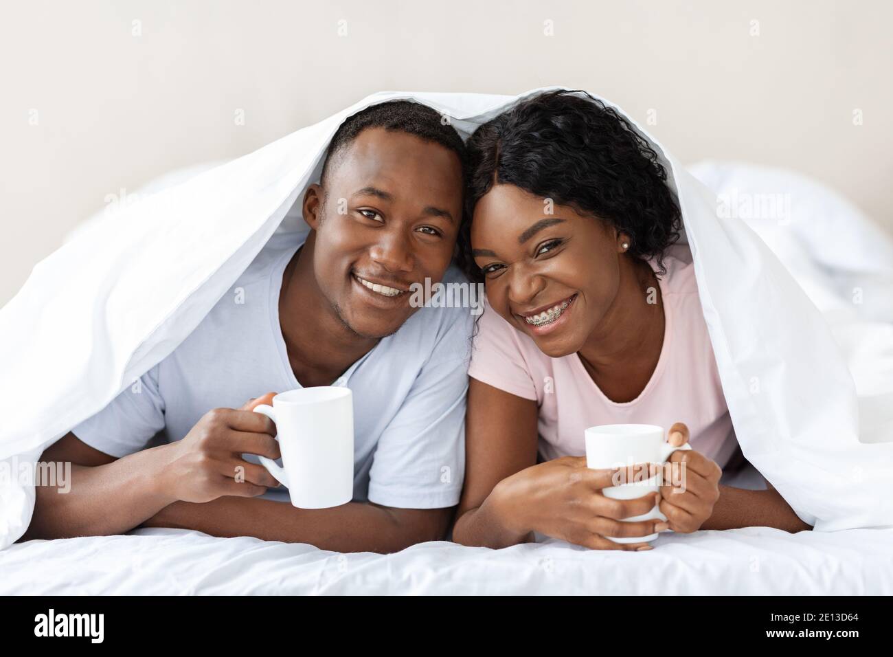 Happy black couple laying under blanket, drinking tea Stock Photo