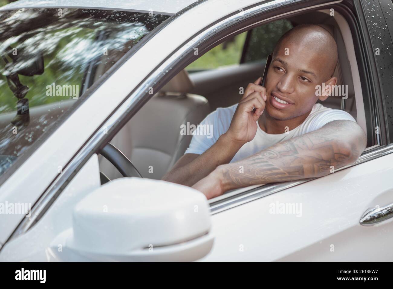 Handsome cheerful African man talking on his smart phone, sitting in a car on a countryside road. Attractive male driver taking break from driving, us Stock Photo
