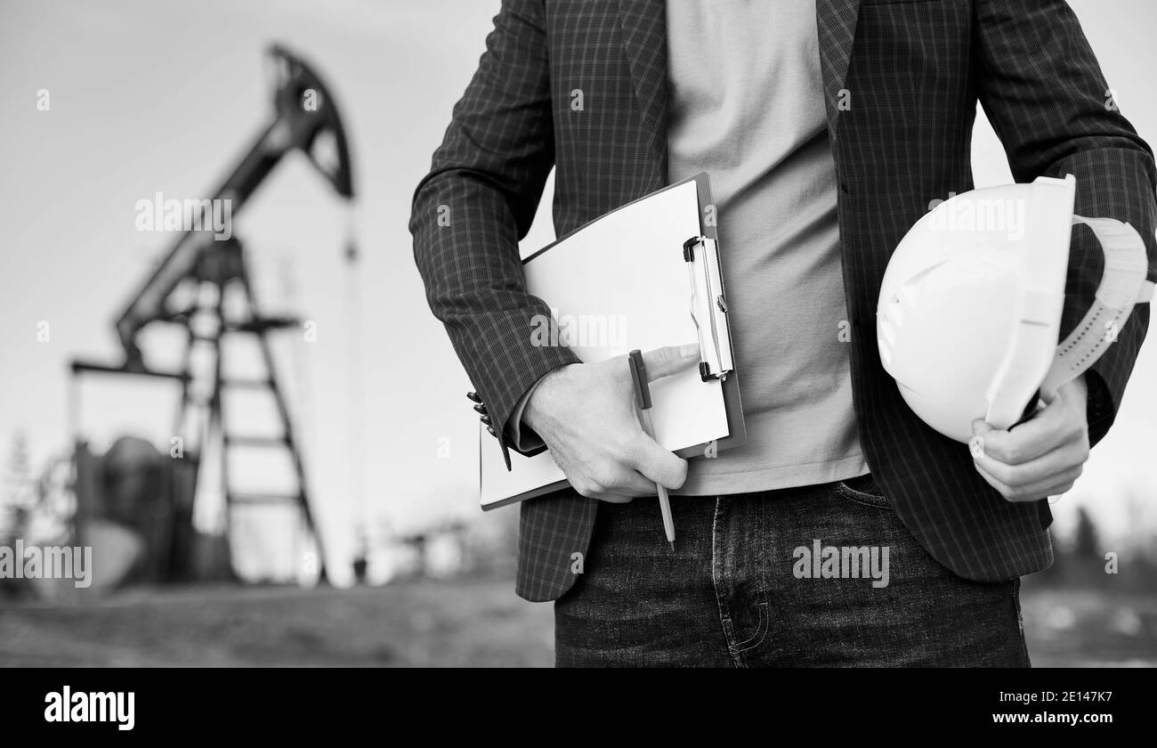 Cropped close-up snapshot of a man engineer holding his note and a pen in one hand and a white helmet in the other, standing in oil field, oil pump jack is on background. Black and white image Stock Photo