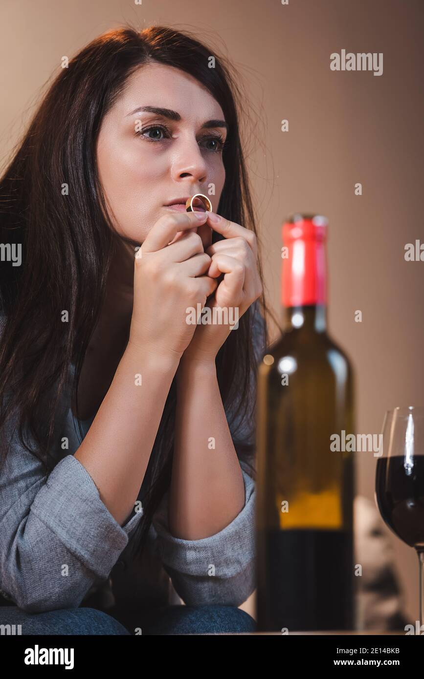 frustrated woman holding wedding ring and looking away near bottle and glass of red wine on blurred foreground Stock Photo