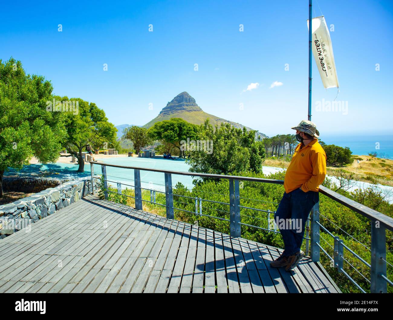 Signal Hill - Cape Town, South Africa -  02/12/2020 Man wearing yellow top and face mask, standing against flag pole. Lion's Head, ocean and greenery Stock Photo