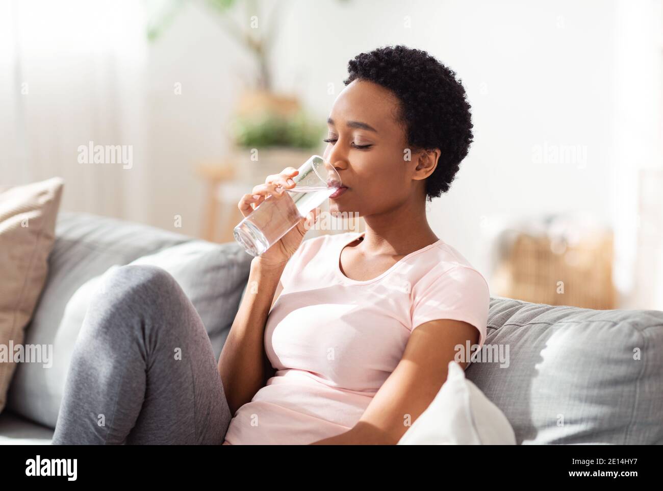 Hydration for good health concept. Pretty young black lady having glass of crystal clear water on couch at home Stock Photo