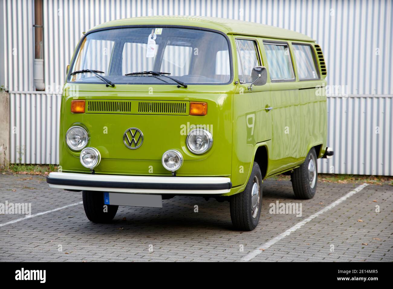 Ein sehr gepflegter Grashuepfer gruener VW Bulli T2 steht auf einem eingezeichneten Parkplatz vor einer weissen Wellblechwand. Stock Photo