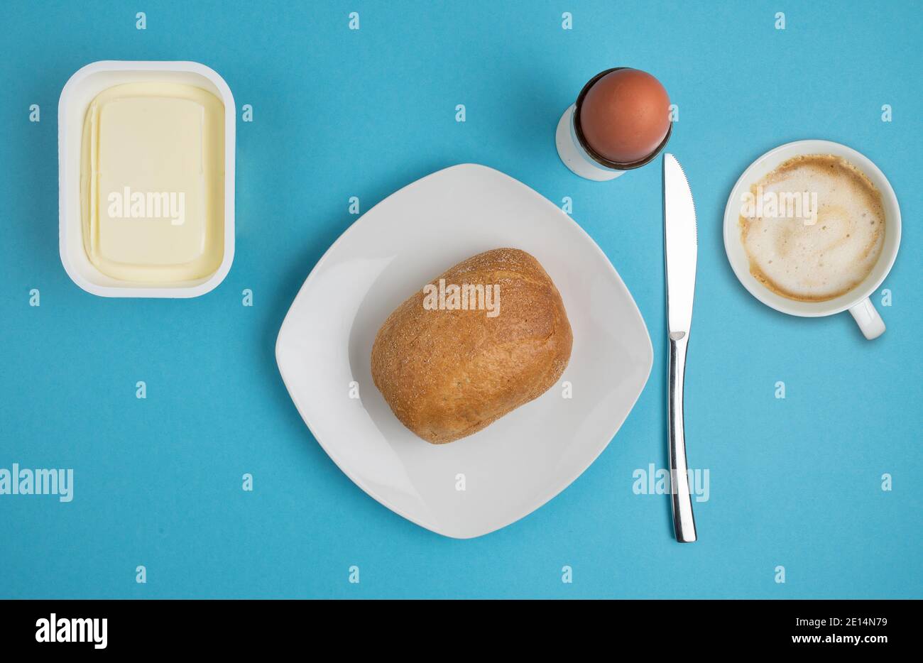 directly above view of bread roll on plate, butter, boiled egg, knife and cup of coffee on blue background, breakfast concept Stock Photo