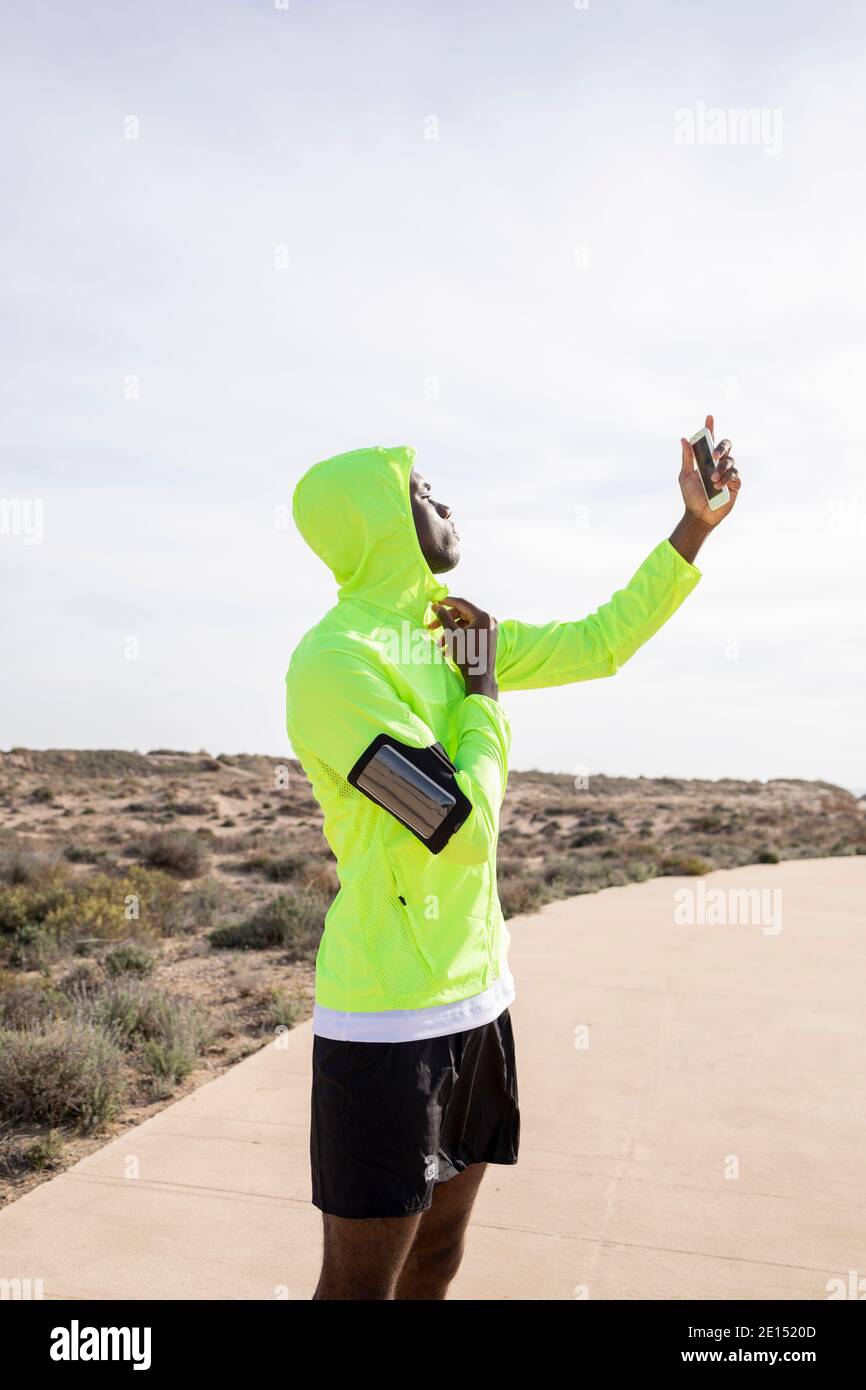 Vertical shot of young black runner with yellow jacket and armband who makes a selfie with his cell phone while he adjusts the zipper of his jacket an Stock Photo