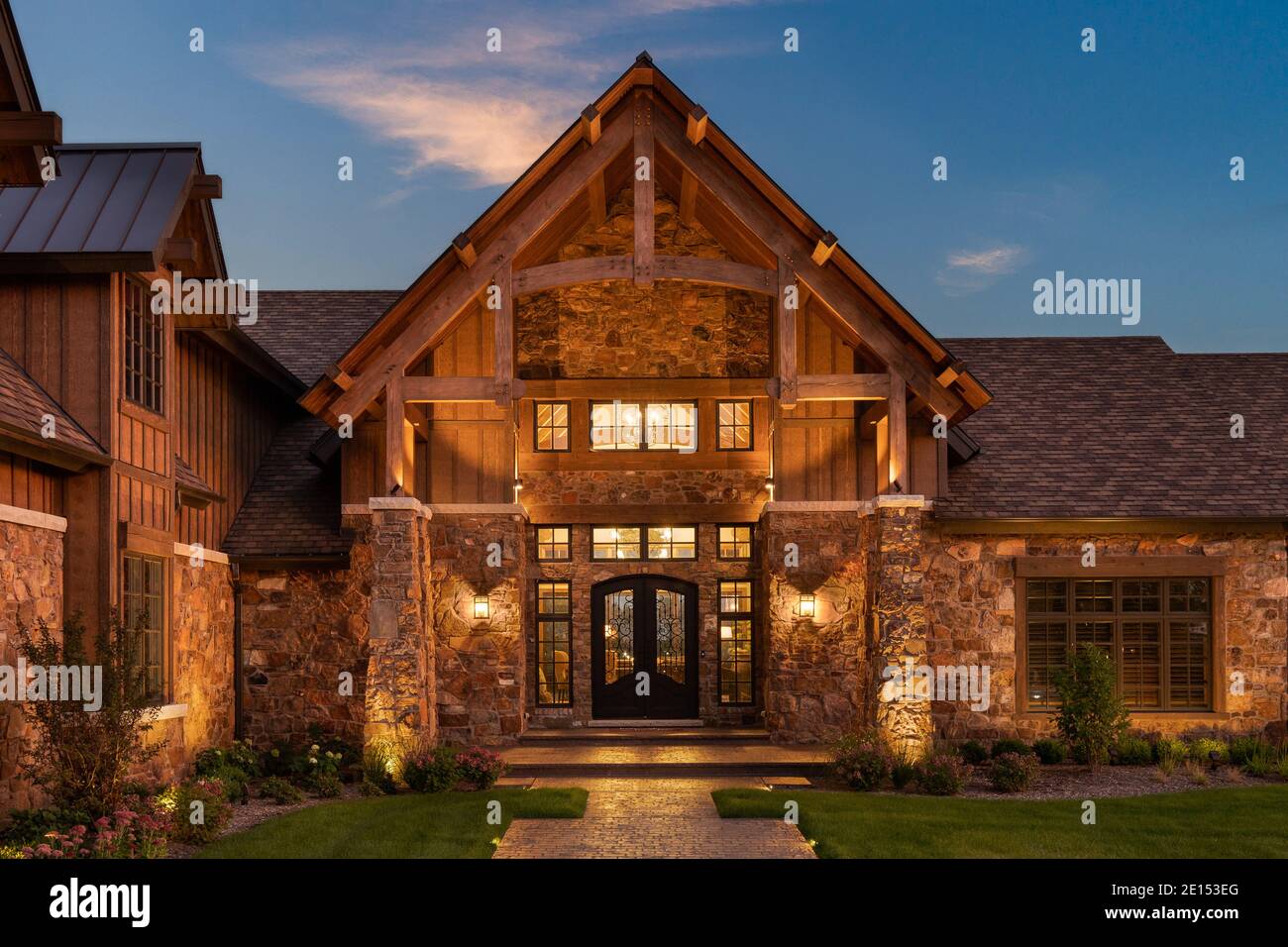 A luxury home with a fancy iron door, surrounded by windows and a rock exterior. Warm bulbs light the outside and inside of the property at sunset. Stock Photo