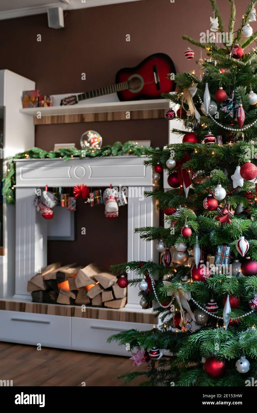 Christmas tree is in the living room is decorated in red and white. In the background is electric fireplace in white cabinet. Stock Photo