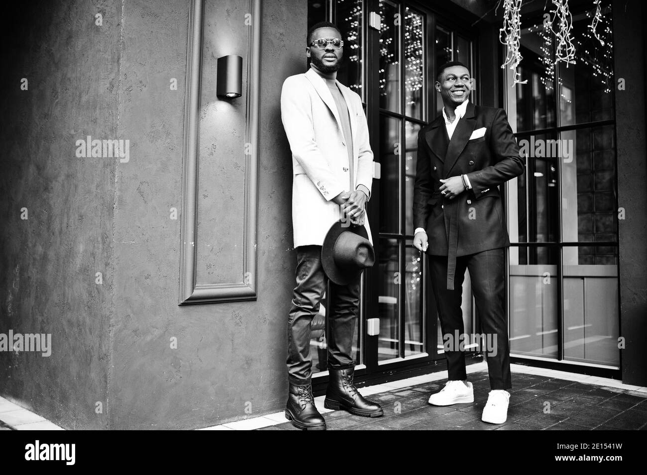 Two fashion black men pose against house with garlands. Fashionable portrait of african american male models. Wear suit, coat and hat. Stock Photo