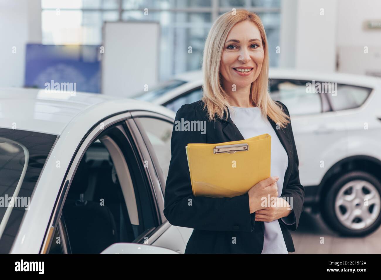 Professional car dealer posing proudly at auto showroom, smiling to the camera, holding clipboard. Happy saleswoman working at automobile dealership, Stock Photo