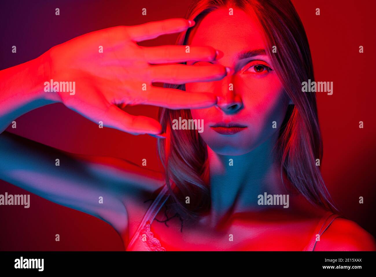 Unemotional young female covering eyes with hand in studio with red light Stock Photo
